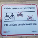 Foto Hinweisschild mit rotem Rahmen und Text: Bitte versperren Sie uns nicht den Weg! Piktogramme eines Rollstuhlfahrenden und eines Menschen mit Gehstock, die sich zwischen zwei Geländern befinden, an denen Fahrräder lehnen, die rot durchgestrichen sind. Text unten: „Keine Fahrräder am Geländer abstellen“. Logo der Deutschen Bahn und der BG Bonn.